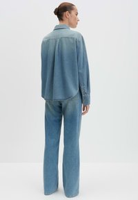 Camicia di jeans blu chiaro con bottone frontale, orlo arrotondato e maniche lunghe. Indossata con pantaloni a gamba larga blu chiaro abbinati.