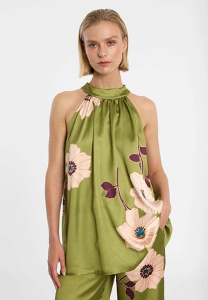 Blonde Frau, die ein ärmelloses olivgrünes Neckholder-Top und passende Hosen mit großen blassrosa und violetten Blumenmustern trägt.