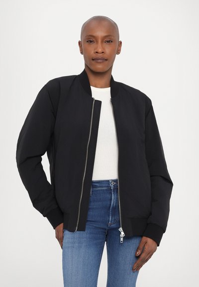 Zwarte bomberjack met ritssluiting, geribde manchetten en een soepele textuur. Draagt over een witte geribde shirt en blauwe jeans.