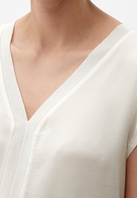 Blůza s véčkovým výstřihem v jemné krémové barvě, vyrobená z hladkého materiálu. Má krátké rukávy a texturovaný výstřih s jemným leskem.