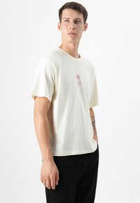 Young man in a light yellow t-shirt with a small pink rose design, standing against a plain white background, looking slightly to the side.