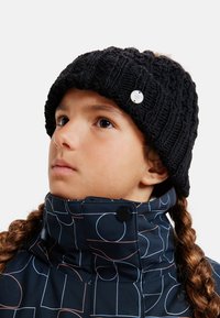 Czarna dziana czapka beanie z rypsowym wzorem i małym metalicznym logo. Osoba nosi wzorzystą, granatową kurtkę z wysokim kołnierzem.