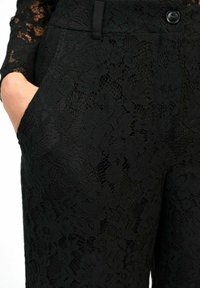 Pantalons en dentelle noire avec motif floral, fermeture à bouton, passants de ceinture et une main dans la poche avant.