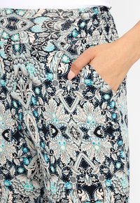 Platas piegriezuma rakstainas bikses melnā, krēma un tirkīza krāsā ar sarežģītu ziedu dizainu; ir sānu kabata un elastīgs jostasvietas aksesuārs.