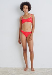 Rdeči bikini set z majhnimi belimi vzorci sidra. Zgornji del ima tanke nastavljive naramnice, medtem ko ima spodnji del nizek kroj. Gladka tekstura.
