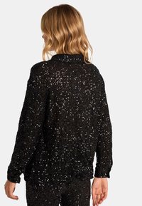 Suéter negro brillante con un corte holgado, mangas largas y tejido texturizado con lentejuelas que añaden un brillo extra. La vista posterior muestra el diseño del cuello.