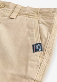 Pantaloni in cotone beige con tessuto testurizzato, dotati di chiusura a bottone e un'etichetta blu navy con la scritta "SUPER DRY" in accenti rossi e bianchi.