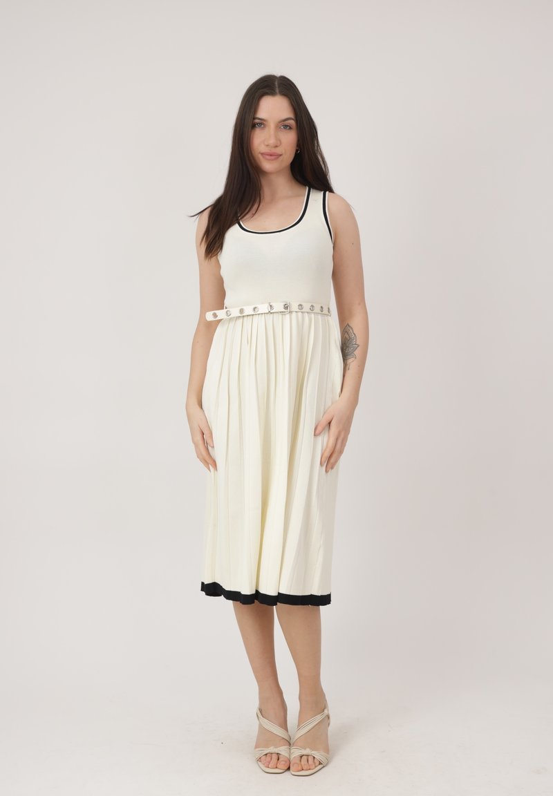 Femme aux longs cheveux noirs portant une robe blanche sans manches avec bordure noire, ceinture boutonnée, et des talons ouverts beige, debout devant un fond uni.
