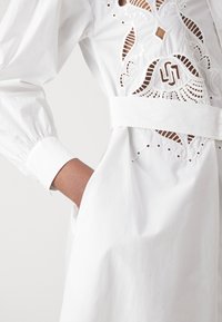 Abito in cotone bianco con intricati tagli a vivo e ricami. Include una cintura larga e maniche a sbuffo per maggior dettaglio.