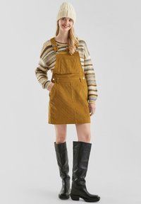 Corduroy mosterdkleurige overall jurk met verstelbare bandjes, voorzak, gecombineerd met een gestreept trui en kniehoge zwarte laarzen.