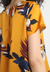 Hořčicově žlutá blůza s krátkými rukávy, zdobená velkými květinovými vzory v modré, červené a bílé barvě. Hladký kulatý výstřih s knoflíkovým zapínáním na zádech.
