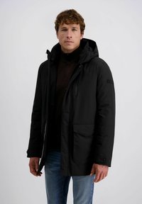 Zwarte geïsoleerde jas met een capuchon, voormet rits en drukknoopsluiting. Heeft twee zijzakken en een hoge kraag, gedragen over een donkere trui.