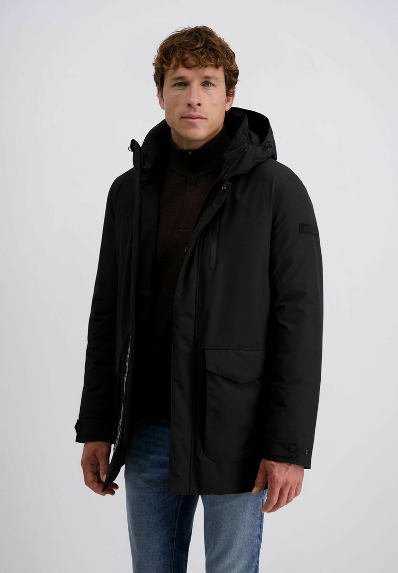 Zwarte geïsoleerde jas met een capuchon, voormet rits en drukknoopsluiting. Heeft twee zijzakken en een hoge kraag, gedragen over een donkere trui.