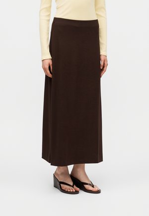 Femme portant une longue jupe marron foncé, un haut beige clair à manches longues et des sandales compensées noires avec des lanières entre les orteils, debout sur un fond uni.