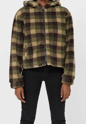 Person trägt einen grün-braun karierten Fleece-Hoodie mit Frontreißverschluss und schwarze Hose und steht vor einem einfarbigen Hintergrund.