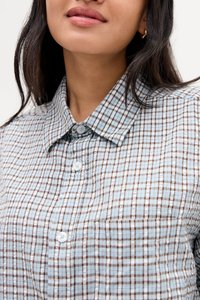 Femme portant une chemise à carreaux bleue et marron avec col et boutons, montrant ses lèvres, son cou et une partie de ses longs cheveux foncés.