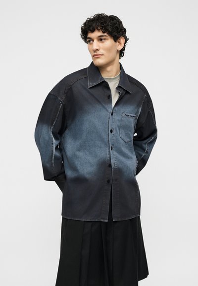 Νεαρό άτομο με σγουρά μαλλιά, που φορά μαύρο και γκρι πουκάμισο από τζιν με κουμπιά και μαύρη πλισέ φούστα, κοιτώντας πλάγια με τα χέρια πίσω από την πλάτη.