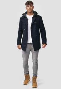 Marineblauwe peacoat met dubbele rij knopen en een gestructureerde capuchon, gecombineerd met grijze jeans en beige laarzen.