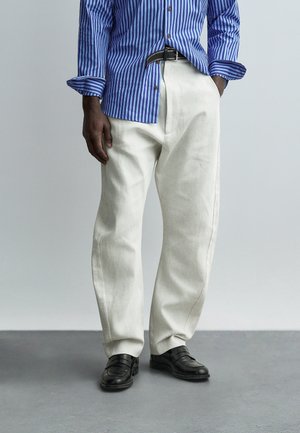 Uomo che indossa pantaloni bianchi larghi, cintura nera, scarpe di pelle nere e camicia a maniche lunghe a righe blu, con una mano in tasca.