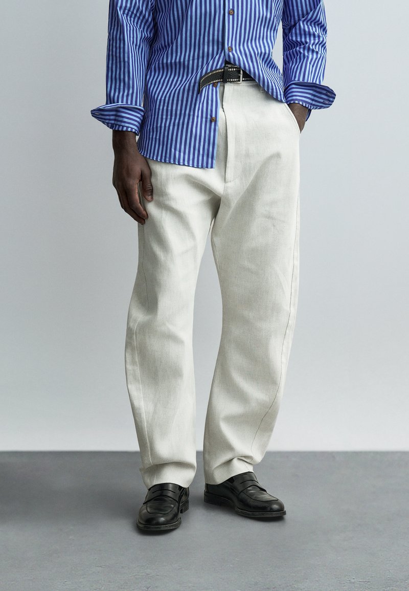 Άνδρας που φοράει λευκό παντελόνι φαρδύ, μαύρη ζώνη, μαύρα δερμάτινα παπούτσια και μπλε ριγέ πουκάμισο με μακριά μανίκια, με το ένα χέρι στην τσέπη.