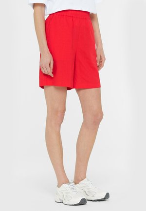 Frau trägt knallrote Shorts mit elastischem Bund und weiße Turnschuhe, steht vor einem schlichten hellen Hintergrund.