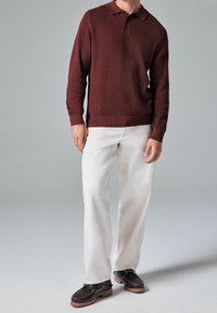 Polo en tricot à côtes rouge foncé, pantalon droit blanc et chaussures à lacets marron avec semelles texturées. Fond gris uni.