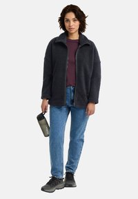 Donkergrijze fleecejack met een ritssluiting aan de voorkant, hoge kraag en een losse pasvorm, gecombineerd met een bordeaux shirt, blauwe spijkerbroek en zwarte schoenen.