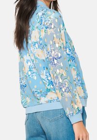 Květinová bomber bunda světle modré barvy, zdobená zlatými metalickými puntíky, s žebrovanými manžetami a volným střihem, vyrobená z poloprůhledné látky.