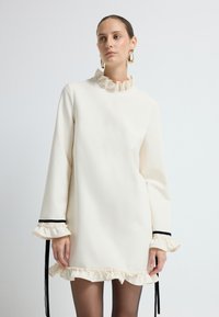 Abito crema con colletto e polsini a volant, caratterizzato da dettagli in elastico nero. Texture liscia, maniche lunghe e design dalla vestibilità comoda.