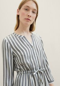 Robe chemise rayée grise et blanche avec un col en V boutonné, des manches longues et une taille cintrée reliée par une ceinture assortie. Texture de tissu lisse.