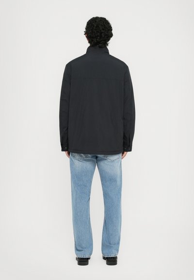 Uomo con capelli ricci che indossa una giacca nera, jeans azzurri chiari e scarpe nere, in piedi di spalle di fronte a uno sfondo bianco.