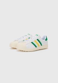 White sneakers with green and yellow stripes, rubber toe cap, lace-up design, and textured sole. Brand label on tongue, green accent at heel.