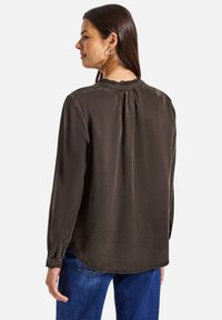 Blouse à manches longues de couleur olive foncé avec un col rond, un dos plissé et des poignets à boutons. Le tissu semble lisse et légèrement brillant.