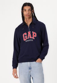 Sudadera de cuello redondo en azul marino con media cremallera, presenta un gran gráfico en rojo y blanco que dice "GAP ATHLETIC" en el frente, hecha de una tela suave.