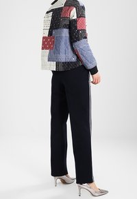 Veste matelassée au design patchwork avec des motifs géométriques rouge, bleu et noir, associée à un pantalon noir et des talons argentés.