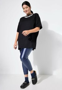 Zwarte oversized t-shirt met de tekst "STUDIO WATT", gecombineerd met glanzende blauwe leggings en zwarte klompen. Accessoires zijn ringen en gestreepte sokken.