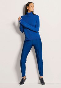 Blaues Rollkragensweater und passende, schlichte Hose mit seitlichen Nähten. Das Modell trägt schwarze Sneaker. Glatte Stricktextur, figurbetonte Silhouette.