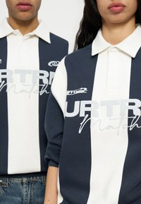 Deux personnes portant des maillots de rugby à rayures verticales bleu marine et blanches avec les logos "URTR Match" et "atomic", cadrées au niveau du cou et de la taille.