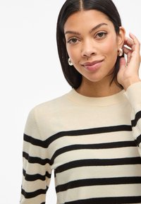 Beige and black striped sweater made of soft material, featuring a round neckline and long sleeves. Model has straight hair and jewelry accents.