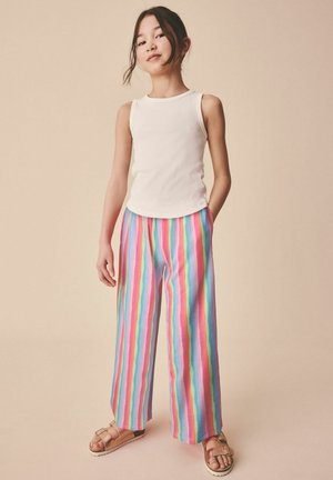 Young girl wearing a white sleeveless top, multicolored vertical striped wide-leg pants, and brown sandals standing against beige background.