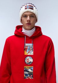 Felpa rossa con stampe grafiche e lacci blu, indossata sopra un top bianco a collo alto. Il berretto presenta un design con la bandiera norvegese.