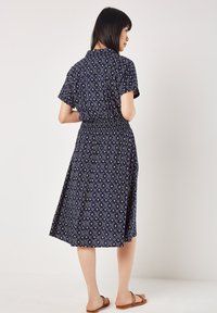 Dunkelblaues Kleid mit kurzen Ärmeln und tailliertem Bund. Es hat ein gemustertes Design und einen knielangen Rock. Wird mit einfachen Sandalen getragen.