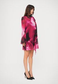 Mulher vestindo um vestido floral curto em rosa e preto de mangas longas e com um laço no pescoço, combinado com sapatos de salto alto pretos, em pé de lado.