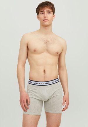 Graue Boxershorts mit einem schwarz-weißen Logo-Bund, enganliegendem Design und glatter Textur. Das Model steht mit den Händen an den Seiten.