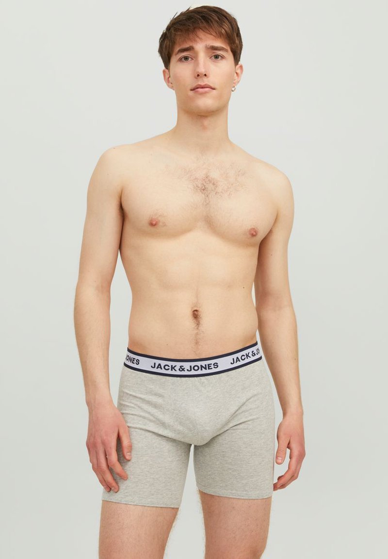 Graue Boxershorts mit einem schwarz-weißen Logo-Bund, enganliegendem Design und glatter Textur. Das Model steht mit den Händen an den Seiten.