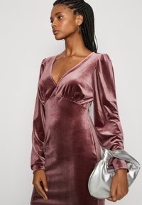 Svilena midi haljina u boji mauve s dugim, napufanim rukavima i dubokim V-izrezom. Uparena s metalik srebrnom clutch torbicom. Glatka tekstura kroz cijelu haljinu.
