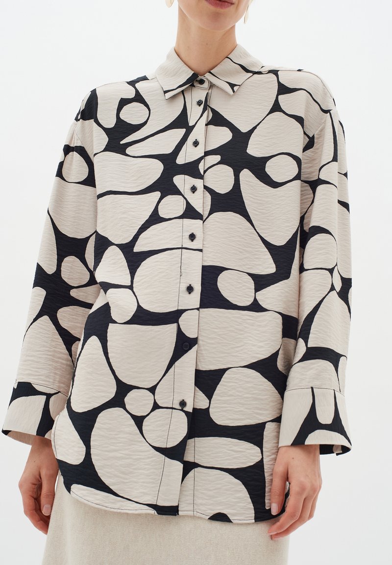 Kreemivärvi nööpidega särk julgete mustade abstraktsete mustritega, millel on krae ja pikad varrukad lõdva mansetiga. Tekstuuriga kangas.