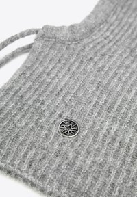 Indumento in maglia grigia con trama a coste verticali, caratterizzato da un emblema rotondo argentato sul davanti. Materiale morbido con scollo rilassato.