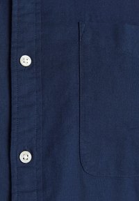 Gros plan sur le devant d’une chemise en tissu bleu marine texturé avec des boutons blancs et une poche poitrine sur le côté droit.