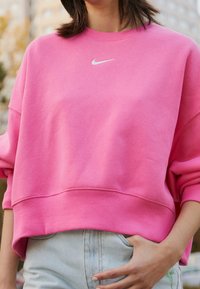 Osoba v jasně růžovém svetru Nike a světle modrých džínech, ruka pohodlně v kapse, venku s rozmazaným městským pozadím.